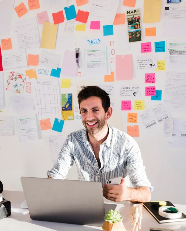 Smiling marketer at a laptop in front of a wall of sticky notes and plans