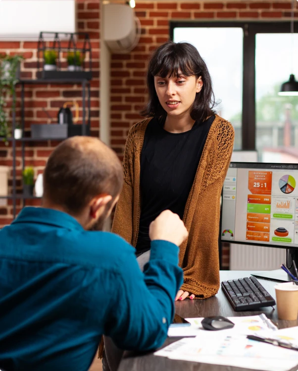 Woman talking with a colleague at a desk beside a monitor showing a dashboard