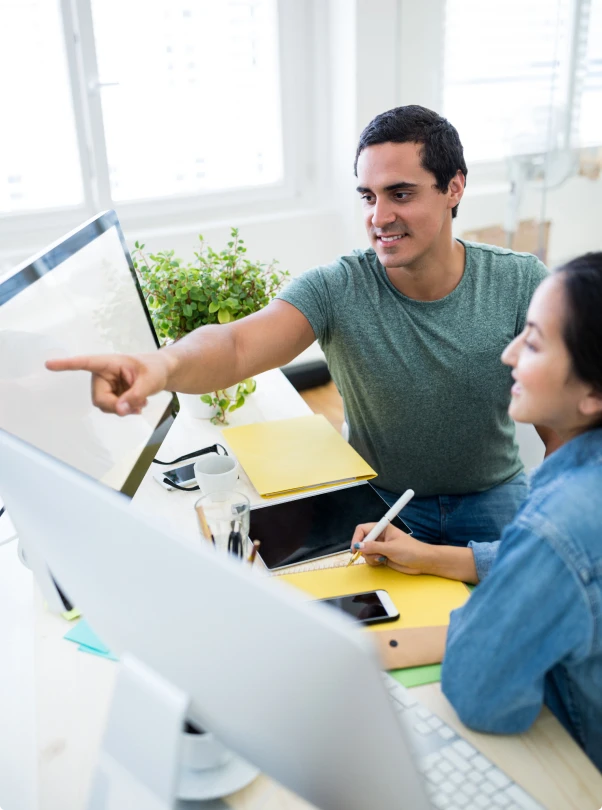 Man pointing at a monitor while a colleague takes notes at a desk