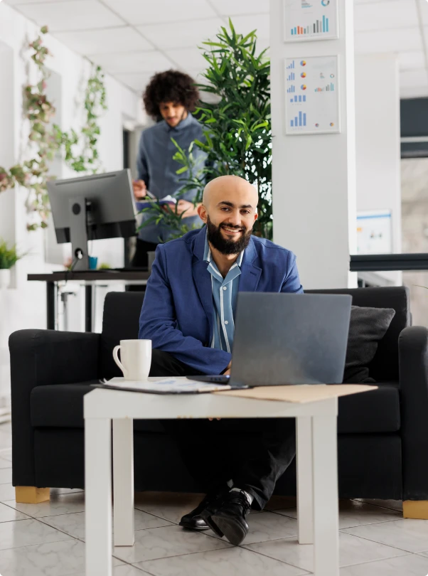 Smiling professional working on a laptop in an office, with a colleague behind him