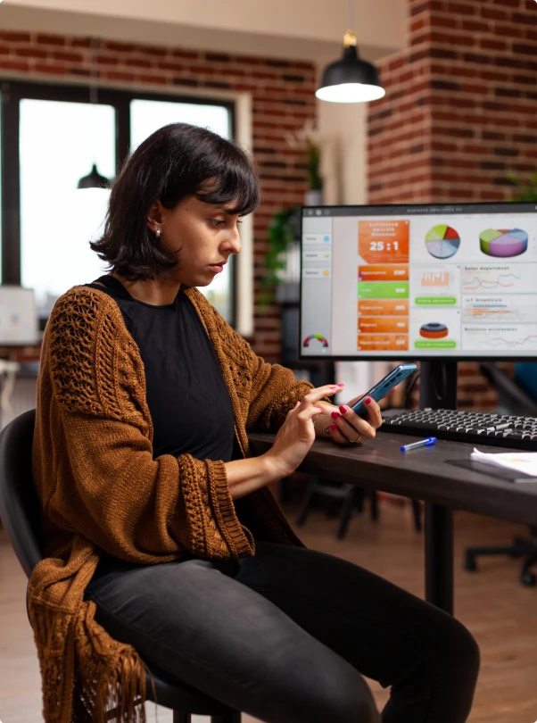 Marketer checking a campaign on her phone beside a dashboard monitor