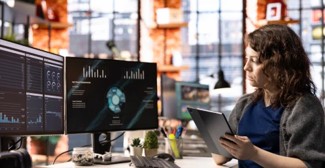 Analyst holding a tablet beside monitors showing analytics dashboards