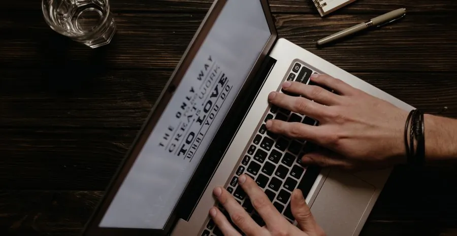 Hands typing an article on a laptop at a wooden desk