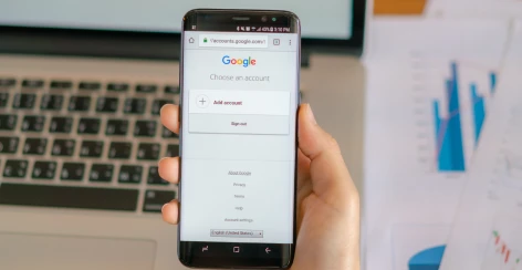 Hand holding a phone showing a Google account sign-in screen in front of a laptop