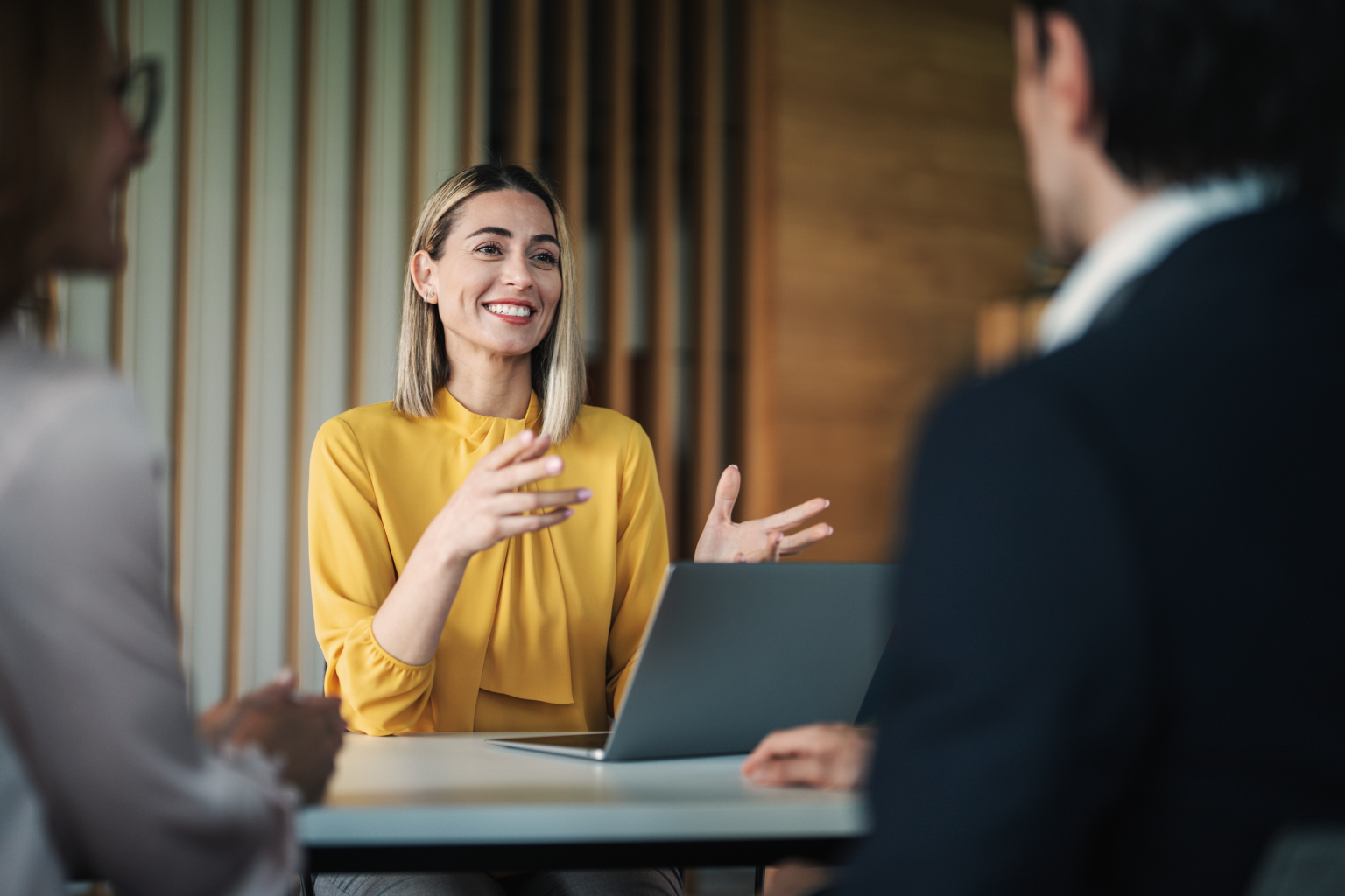 A woman in a yellow blouse speaks with two colleagues across a table with a laptop during a business meeting
